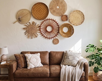 May include: A brown leather sofa with three decorative pillows and a beige throw blanket, positioned below a wall arrangement of nine circular woven baskets and fans in various shades of tan, brown, and natural wood tones.