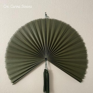 May include: A large, semi-circular wall fan in a deep olive green color, featuring fine, radiating pleats that extend from a central point. Two matching olive green tassels hang vertically from the center, suspended by a thin cord.