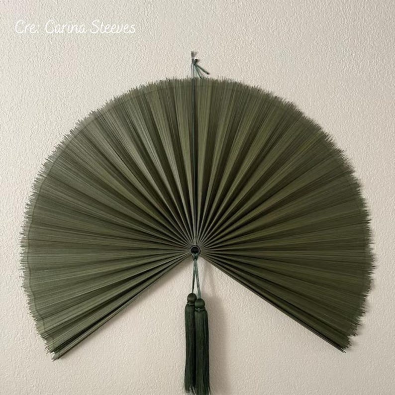 May include: A large, semi-circular wall fan in a deep olive green color, featuring fine, radiating pleats that extend from a central point. Two matching olive green tassels hang vertically from the center, suspended by a thin cord.