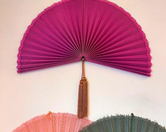 May include: A large, vibrant magenta pleated hand fan with a textured surface and a fringed edge. A brown cord with two matching brown tassels hangs from the center pivot point. Below, partially visible, are two additional fans in light pink and sage green.