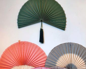 May include: A collection of semi-circular pleated wall fans in various colors including olive green, coral, and grey. The green fan features a dark green tassel hanging from the center. Each fan displays a fine, ribbed texture.