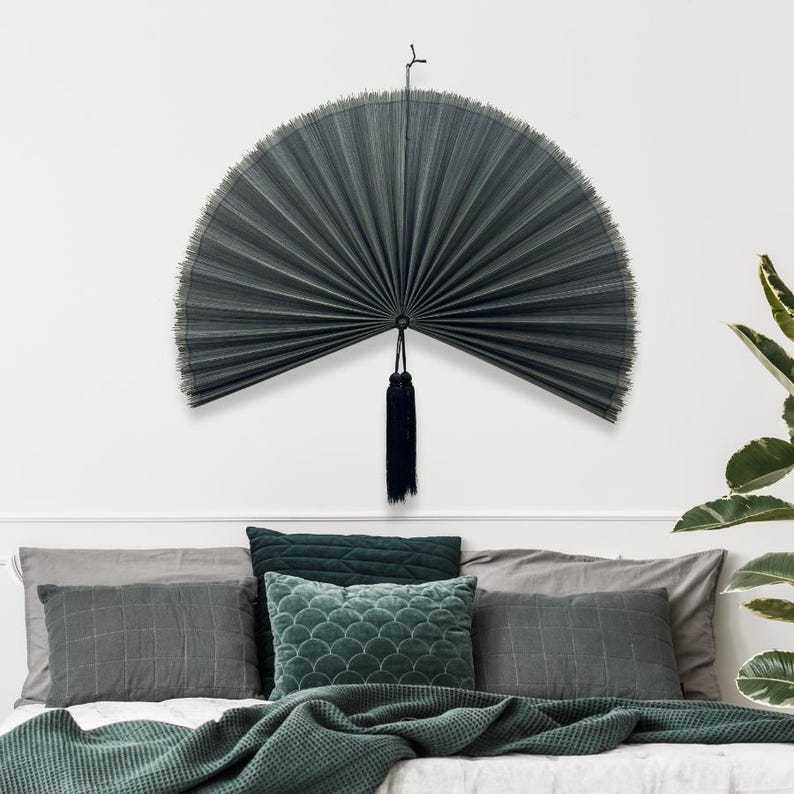 May include: A large, semi-circular dark gray pleated wall fan featuring a fine, fringed edge and a long, dark hanging tassel at the center. The fan is mounted on a wall, displaying a structured, accordion-style design with thin, radiating ribs.