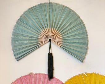 May include: A large, semi-circular wall fan featuring light blue and white pleated ribs radiating from a central point. A dark green cord extends from the top, and a long, dark green tassel hangs from the center pivot point of the fan.