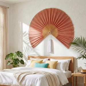 Puede incluir: Un gran adorno de pared semicircular en tonos coral y dorado, que se asemeja a un abanico. Está montado sobre una cama con sábanas blancas y almohadas coloridas. Unas borlas blancas cuelgan del centro del abanico.
