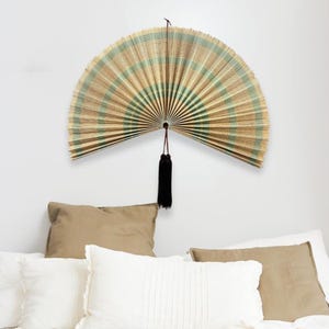 May include: A large, semi-circular wall fan made of woven natural fibers in alternating stripes of light tan and sage green. The fan features a pleated design and is suspended by a thin cord with a long, dark brown decorative tassel hanging from the center.