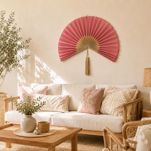 May include: A large, semi-circular wall fan featuring pleated pink fabric with a gold-toned center and a decorative tassel hanging from the bottom. The fan is mounted on a plain wall above a sofa with various textured throw pillows and a wooden coffee table.