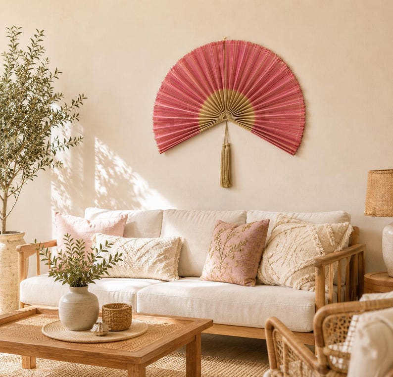 May include: A large, semi-circular wall fan featuring pleated pink fabric with a gold-toned center and a decorative tassel hanging from the bottom. The fan is mounted on a plain wall above a sofa with various textured throw pillows and a wooden coffee table.