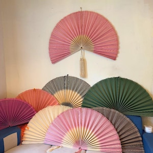 May include: A collection of large, semi-circular pleated fans in various colors including pink, green, beige, and brown. One pink fan is mounted on the wall, while others are arranged on a surface. Each fan features a central pivot point.
