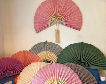 May include: A collection of large, semi-circular pleated fans in various colors including pink, green, beige, and brown. One pink fan is mounted on the wall, while others are arranged on a surface. Each fan features a central pivot point.