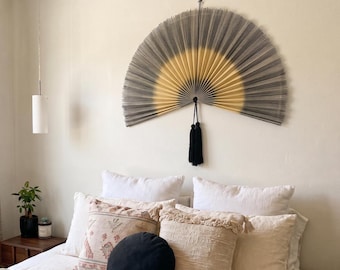 May include: A large, semi-circular wall fan featuring a gradient design that transitions from a dark gray outer edge to a golden yellow center. The fan is suspended by a black cord and includes a decorative black tassel hanging from the center point.