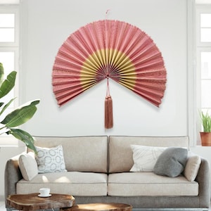May include: A large, semi-circular wall fan in shades of pink and gold, with a tassel hanging from the center. The fan is mounted on a white wall above a beige sofa, creating a decorative accent. The fan's design adds a touch of elegance to the room.