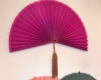 May include: A large, semi-circular fuchsia pink pleated wall fan with a textured, fringed edge. The fan features a central pivot point with a long, thin brown cord for hanging and a decorative brown tassel hanging from the center base.