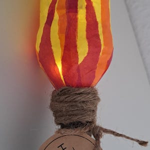 Könnte beinhalten: Eine handgefertigte Papierlaterne in Form einer Flamme mit gelber und orangefarbener Basis und roten und orangen Flammen. Die Laterne ist mit Schnur an einem braunen Stock befestigt und trägt ein braunes Etikett mit der Aufschrift "HANDMADE WITH LOVE".
