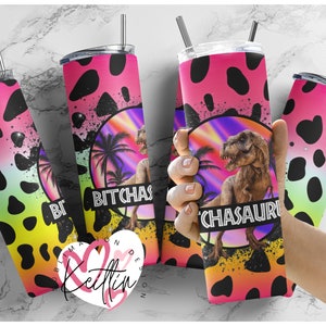 May include: A pink and black stainless steel tumbler with a graphic of a dinosaur in a tropical setting. The text "BITCHASAUR" is printed on the tumbler.