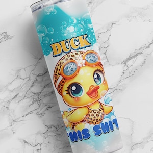 Puede incluir: Un vaso blanco con fondo azul y un pato de dibujos animados que lleva un traje de baño de estampado de leopardo y gafas de buceo. El texto "DUCK THIS SHIT" está escrito en letras azules en el vaso.