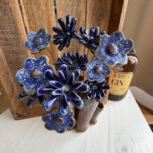 Könnte beinhalten: Eine Keramikskulptur mit mehreren blauen Blumen mit gesprenkelten weißen Details und braunen Mitten, die an dünnen Metallstielen befestigt sind. Die Blumen sind in einer braunen, mehrröhrigen Keramikvase angeordnet.