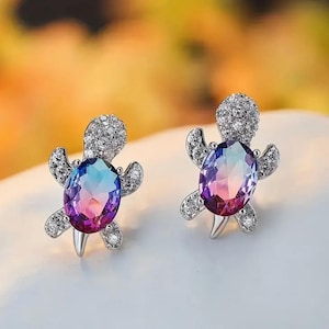 May include: A pair of silver turtle earrings with iridescent purple, blue, and pink gemstones. The earrings are set with clear cubic zirconia stones.