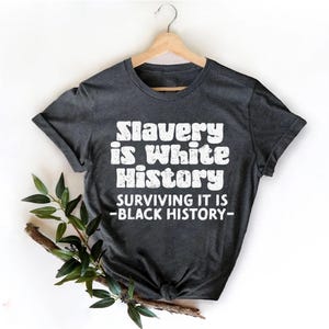 May include: A dark grey t-shirt with the white text "Slavery is White History, Surviving it is - Black History-". The shirt is displayed on a wooden hanger. Greenery is visible in the lower left corner of the image.