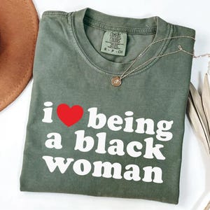 May include: An olive green Comfort Colors t-shirt with the text "i ❤️ being a black woman" in white and a red heart. A gold necklace with a pendant is draped across the shirt. A brown hat is partially visible in the background.