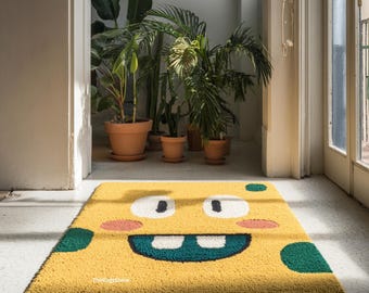 May include: A rectangular yellow rug featuring a cartoon face with large white eyes, black pupils, pink cheeks, and a wide teal mouth with two teeth. The rug has teal circular spots and the text TheRugsShine printed in small white letters.