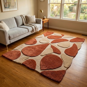 May include: A rectangular area rug featuring a tufted, abstract pattern of organic, teardrop-shaped forms in shades of terracotta orange and cream. The rug has a textured, plush pile surface and is laid flat on a wooden floor in a living room.