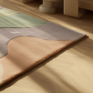 May include: A colorful area rug with an abstract design. The rug features soft, muted tones of peach, gray, green, and cream. The edges of the rug are slightly curved, creating a modern and inviting look. The text "TheRugsShine" is visible on the rug.