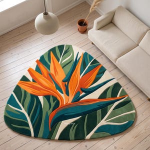 May include: A triangular area rug featuring a large, vibrant orange and teal bird of paradise flower centered among deep green and light cream tropical leaves. The rug has a tufted texture and displays the text The Rugs Shine in small white letters.