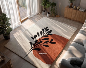 May include: A rectangular area rug with a cream background, featuring a large burnt orange shape and a black botanical design. The rug is placed on a hardwood floor in a living room setting, with natural light.
