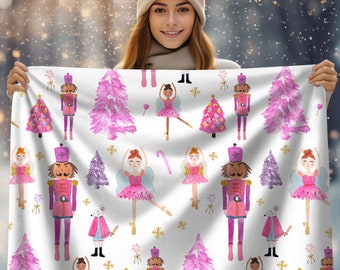 Puede incluir: Una tela blanca con un patrón de acuarela rosa y morado con bailarinas, cascanueces y árboles de Navidad. El patrón se repite en toda la tela.