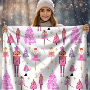 Puede incluir: Una tela blanca con un patrón de acuarela rosa y morado con bailarinas, cascanueces y árboles de Navidad. El patrón se repite en toda la tela.