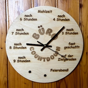 Könnte beinhalten: Eine Holz-Uhr mit schwarzen Zeigern und einem Countdown-Design. Das Zifferblatt der Uhr zeigt die Worte "BÜRO COUNTDOWN" und Sätze wie "noch 6 Stunden" und "Feierabend!"