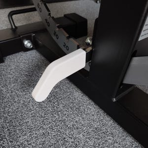May include: Close-up of a weight bench adjustment mechanism. The bench features a black metal frame with a white lever for adjusting the angle. The angle indicator shows markings from 20 to 50 degrees. The background is a gray carpet.