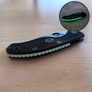 Può includere: Un coltello pieghevole nero con un manico strutturato e un bordo verde brillante. Il coltello ha un design circolare sul manico e una lama argentata. L'immagine include anche un inserto circolare che mostra il coltello che si illumina al buio.