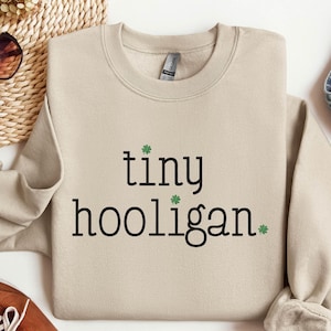 May include: A beige sweatshirt with the text "tiny hooligan" printed in black with green shamrock symbols.