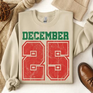 May include: A beige sweatshirt with the text "DECEMBER 25" in green and red. The number 25 has a distressed, vintage look. The sweatshirt is laid out with a beige knit hat, brown leather boots, and a decorative ball.