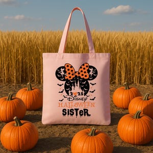 May include: A pale pink tote bag with a Halloween design. It features a Minnie Mouse bow with orange polka dots, bats, and the text "Disney Halloween Sister". Several orange pumpkins are scattered around the bag.