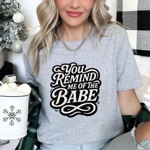 May include: A light gray t-shirt with the text "You Remind Me Of The Babe" in a black and white design. The shirt is being worn by a person. A white mug with a snowflake design is visible on the left.