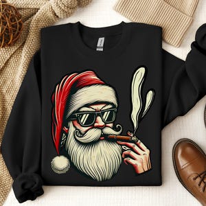 May include: Black sweatshirt featuring a cartoon illustration of Santa Claus wearing sunglasses and smoking a cigar. Santa's hat is red with a white pom-pom. The design has a vintage, retro style.