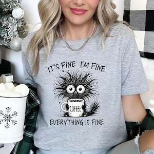 Peut inclure: T-shirt gris clair avec un dessin d'un monstre de dessin animé tenant une tasse de café. Le texte "IT'S FINE I'M FINE EVERYTHING IS FINE" est imprimé au-dessus et en dessous. Le t-shirt est de style décontracté.