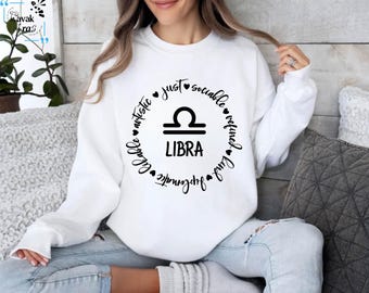 Op de afbeelding: Wit sweatshirt met een zwart cirkelvormig ontwerp met het sterrenbeeld Weegschaal en de woorden "gewoon, sociaal, verfijnd, charmant, lief, artistiek en onweerstaanbaar" rond het symbool. Het woord "LIBRA" staat onder het symbool.