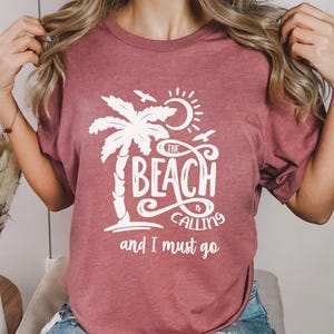 Może przedstawiać: Maronowy t-shirt z białym nadrukiem palmy, słońca i ptaków. Tekst na koszulce brzmi "The Beach is Calling and I Must Go."