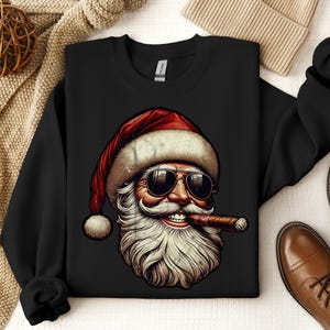 May include: Black crewneck sweatshirt featuring a graphic of Santa Claus wearing sunglasses and smoking a cigar. Santa's hat is red with white trim. The image has a vintage, distressed look. The sweatshirt is displayed with brown leather shoes and a beige beanie.