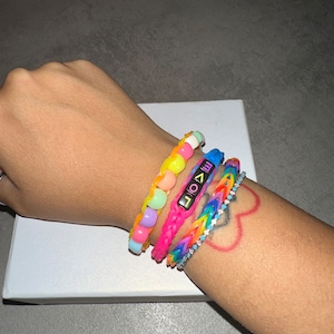 Può includere: Un polso con quattro braccialetti colorati. Il braccialetto superiore è fatto di perline arcobaleno. Il secondo braccialetto è rosa e ha la parola "LOVE" scritta in nero. Il terzo braccialetto è arcobaleno e il braccialetto inferiore è una catena d'argento con pietre trasparenti.
