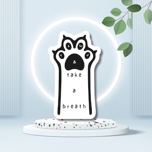 May include: A white sticker in the shape of a paw print with the words "PAWS & take a breath" in black. The sticker is on a white pedestal with a light blue background and a white circle behind the sticker.