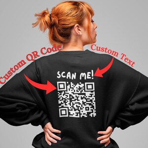 May include: Black sweatshirt with a white QR code and the text "SCAN ME!". The sweatshirt is worn by a person with red hair. The words "Custom QR Code" and "Custom Text" are written in red.