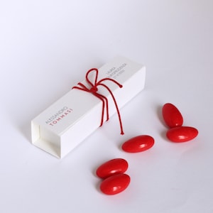 Può includere: Una scatola rettangolare in cartoncino bianco legata con un sottile spago rosso, con il testo stampato ALESSANDRO TOMMASI e LAUREA GIURISPRUDENZA 27 VI 2026. Quattro confetti rossi lisci e ovali sono disposti vicino alla scatola.