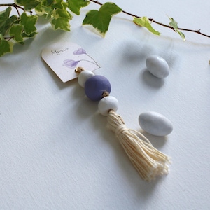 Può includere: Un nappa decorativa con una perla lavanda, perle bianche e una nappa beige. È attaccata una piccola etichetta con il nome "Marco" e un motivo floreale. Sono presenti anche due caramelle bianche a forma di mandorla.