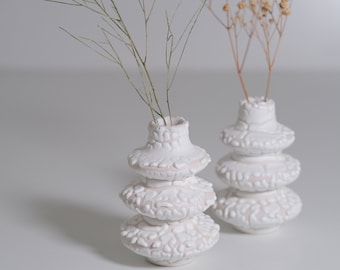 May include: Two white ceramic vases with a textured surface. The vases are stacked in a tiered design. One vase holds a sprig of dried flowers, and the other holds a sprig of green foliage.