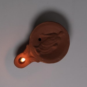 Pode incluir: Uma lamparina de terracota com corpo redondo e bico. A lamparina tem um desenho em relevo na parte superior e um pequeno orifício para o pavio. O pavio está aceso, emitindo um brilho quente. Cor castanho-avermelhado.