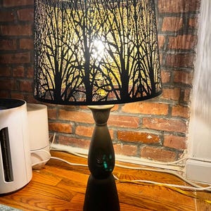 Puede incluir: Una lámpara de mesa negra con una pantalla negra que presenta un diseño recortado de árboles. La pantalla está iluminada, proyectando un brillo cálido.